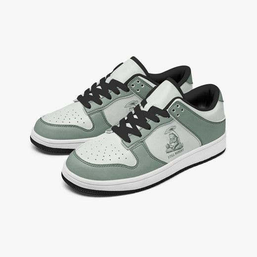 Angled three-quarter view of low-top sneakers in sage green and off-white leather, featuring a frog artwork with "STILL MOMENT", black laces, and a black inner lining.