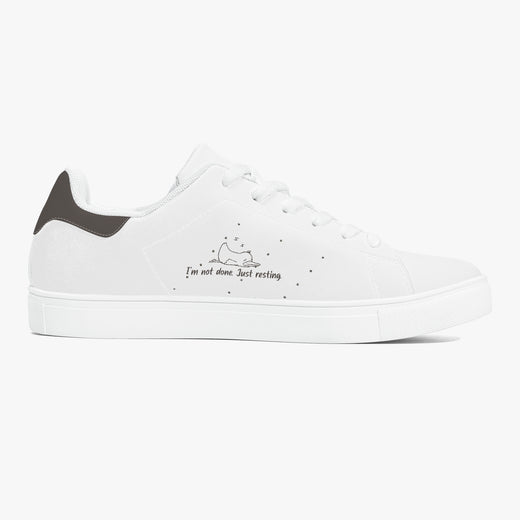A white low-top sneaker with a dark gray heel tab, white laces, and a small black graphic on the side depicting a sleeping cat with the text 'I'm not alone. Just resting'.