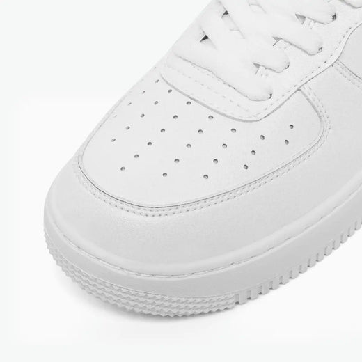 A close-up shot of a white low-top sneaker, showing the toe box with perforations, white laces, and a thick textured white sole on a clean white background.
