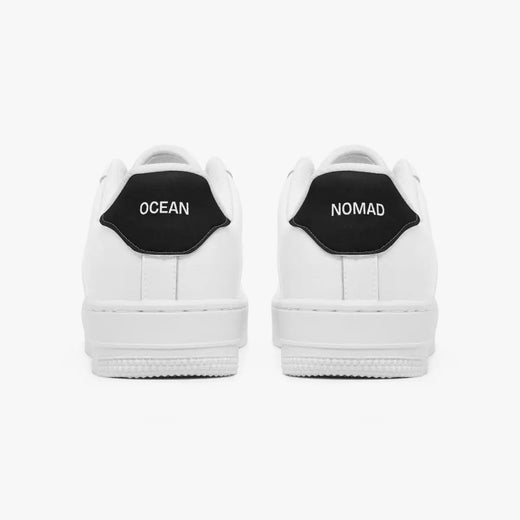 Rear view of white low-top sneakers showing contrasting black heel tabs labeled "OCEAN" and "NOMAD" with flat white rubber soles.