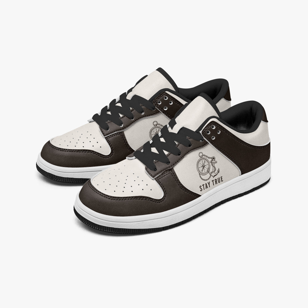 Angled three-quarter view of low-top sneakers in espresso brown and cream leather, featuring an anchor and compass illustration with "STAY TRUE", black laces, and a black inner lining.