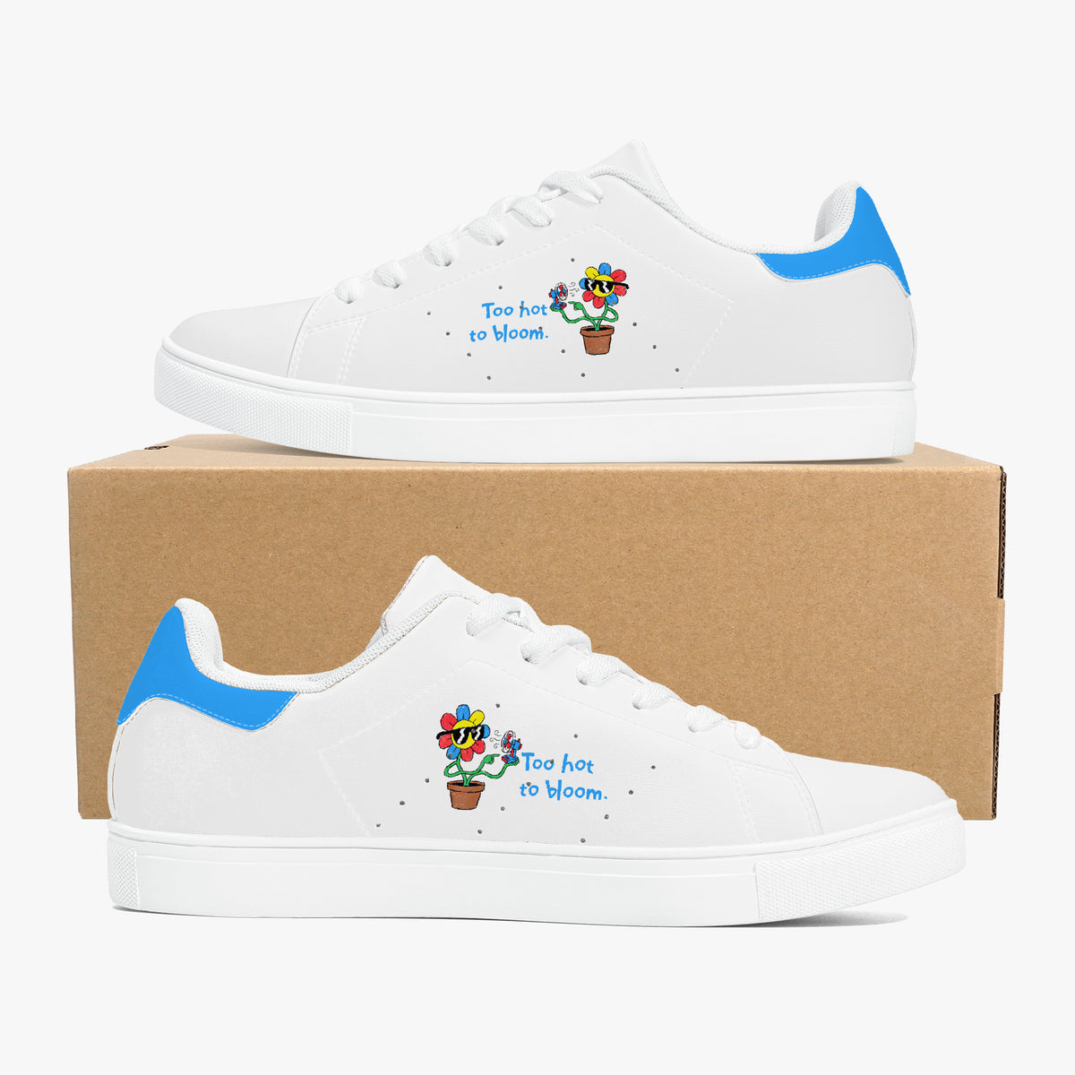 A pair of white low-top sneakers with blue heel accents and a colorful cartoon cactus graphic with the text 'Too hot to bloom' on the side, displayed on a brown cardboard box against a light gray background.