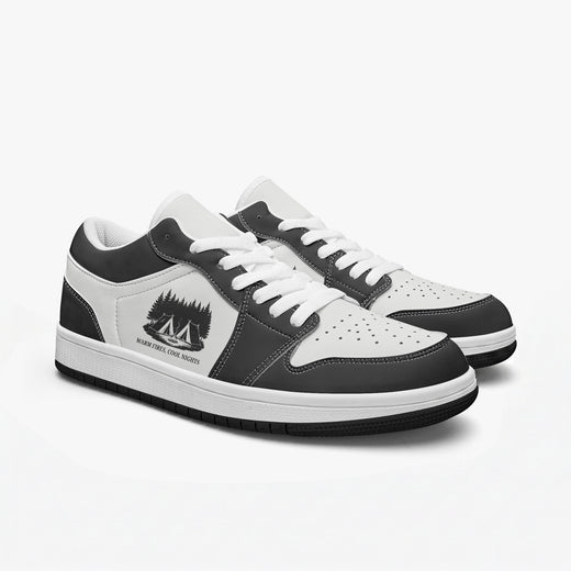 Angled three-quarter view of low-top sneakers in charcoal black and white leather, featuring a campsite in the woods illustration with "WARM FIRES, COOL NIGHTS", white laces, and a white inner lining.