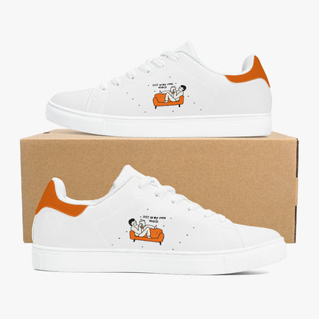 A pair of white low-top sneakers with orange heel accents and a small cartoon graphic on the side, displayed on a brown cardboard box against a light gray background.