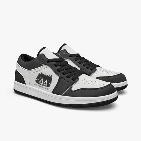 Angled three-quarter view of low-top sneakers in charcoal black and white leather, displaying the "WARM FIRES, COOL NIGHTS" camp graphic, black laces, and a black interior lining.