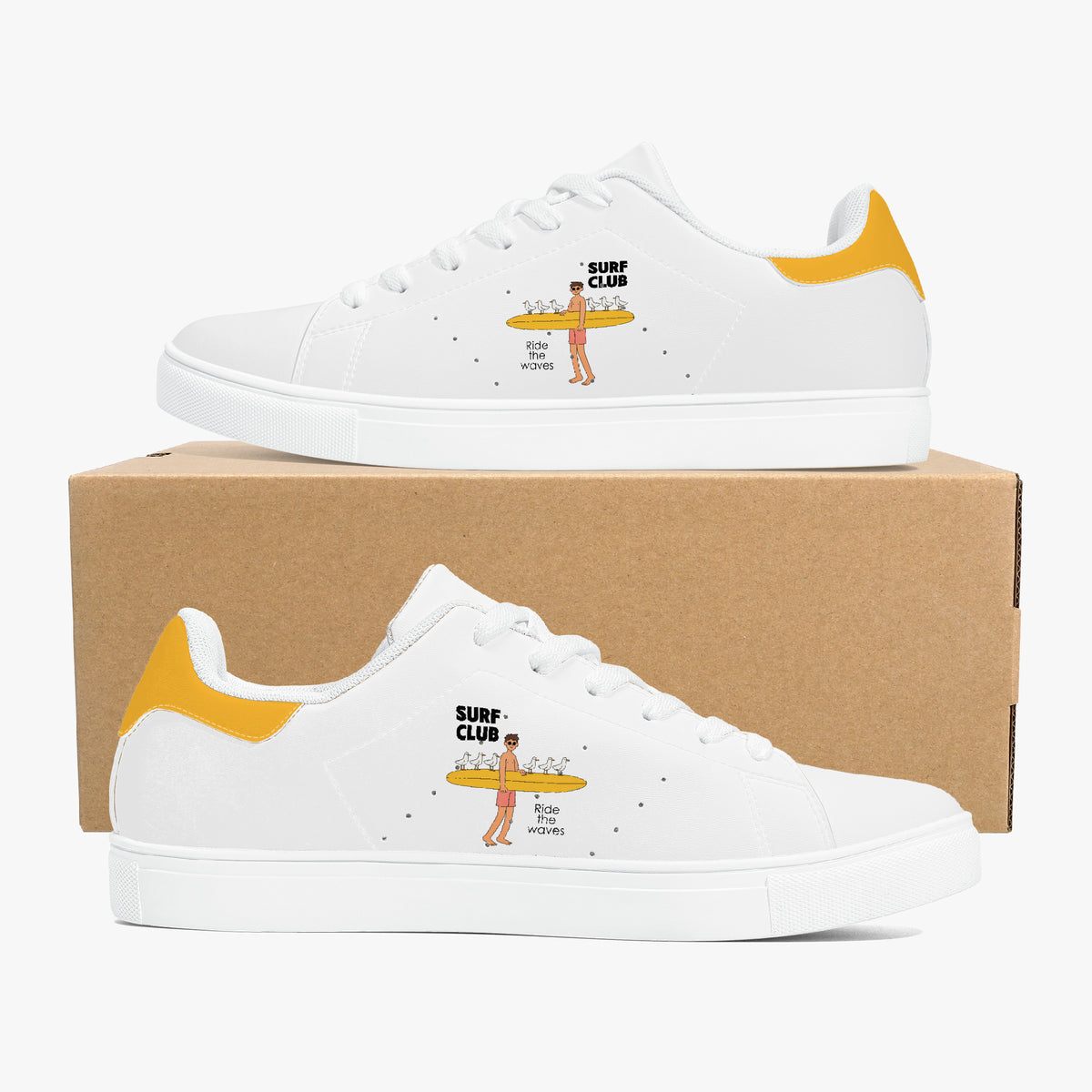 A pair of white low-top sneakers with yellow heel accents and a small surfboard graphic on the side, displayed on top of and in front of a brown cardboard box against a light gray background.