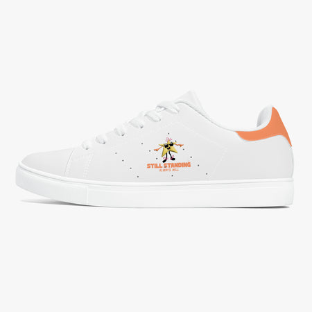 A white low-top sneaker with a white rubber sole, featuring a small graphic of a dancing figure and the text 'STILL STANDING ALWAYS WILL' on the side, with an orange heel accent.