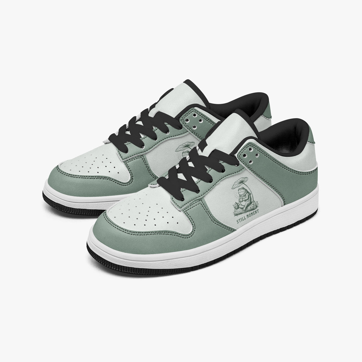 Angled three-quarter view of low-top sneakers in sage green and off-white leather, featuring a frog artwork with "STILL MOMENT", black laces, and a black inner lining.