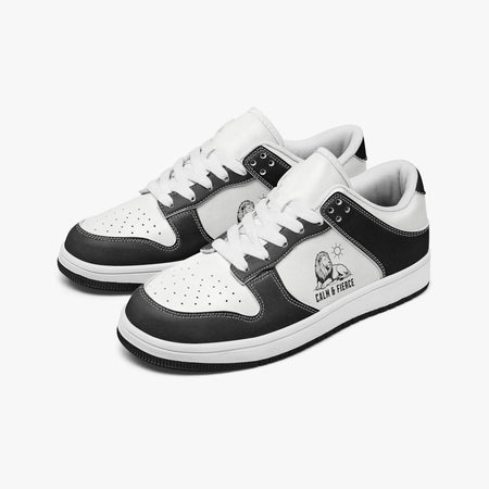 Angled three-quarter view of low-top sneakers in charcoal black and white leather, featuring a resting lion graphic with "CALM & FIERCE", white laces, and a white inner lining.