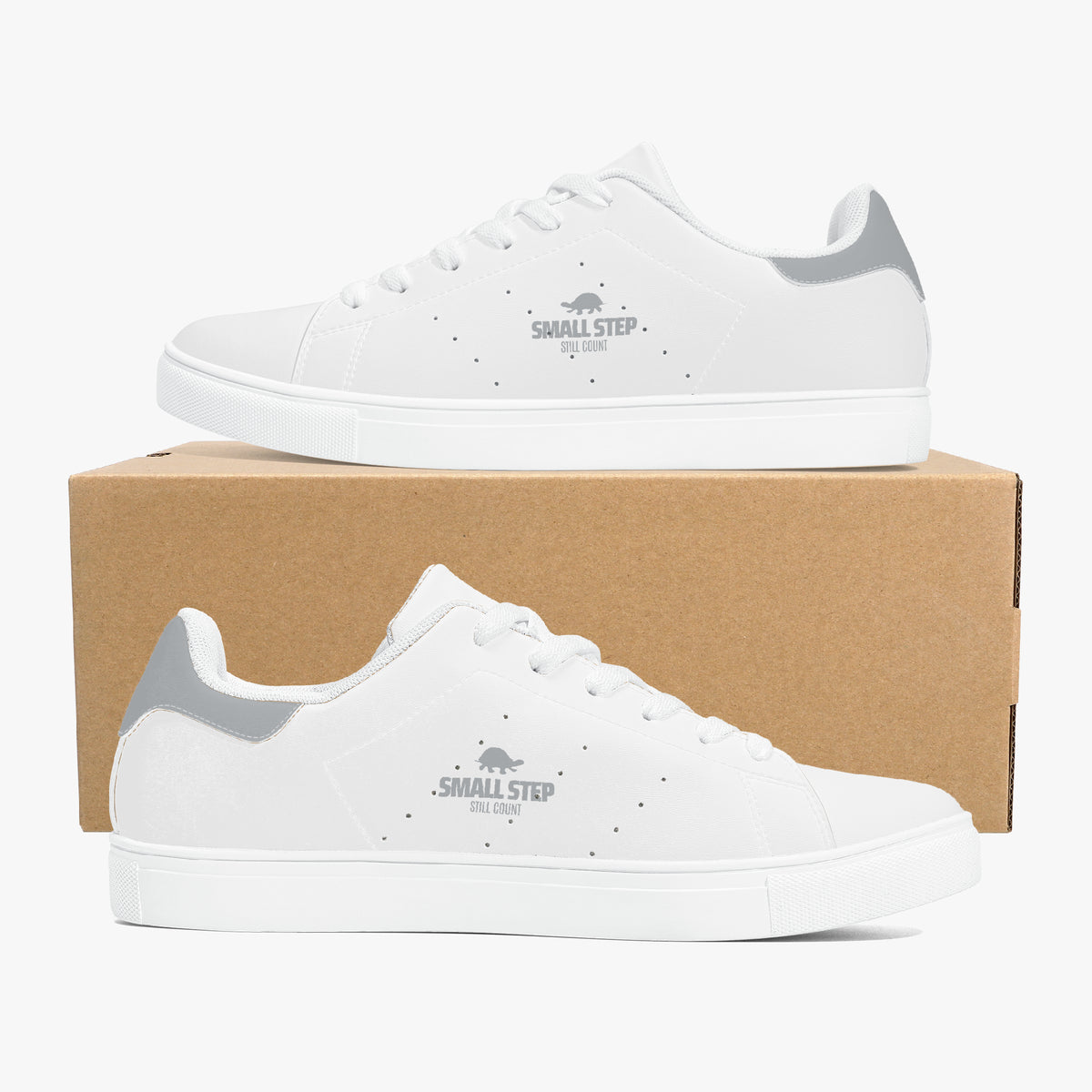 A pair of white low-top sneakers with gray heel accents and a gray printed logo on the side, displayed on a brown cardboard box against a light gray background.
