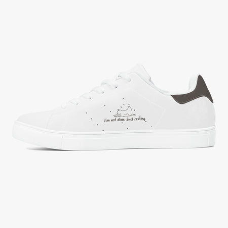 A white low-top sneaker with a black heel tab, featuring a small sleeping cat illustration and the text 'I'm not done. Just resting.
