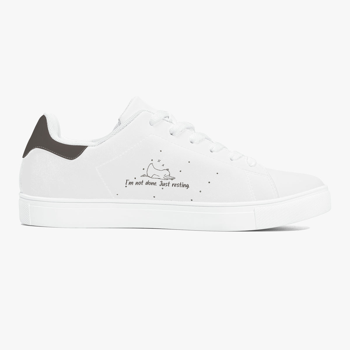 A white low-top sneaker with a dark gray heel tab, white laces, and a small black graphic on the side depicting a sleeping cat with the text 'I'm not alone. Just resting'.