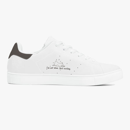 A white low-top sneaker with a dark gray heel tab, white laces, and a small black graphic on the side depicting a sleeping cat with the text 'I'm not alone. Just resting'.