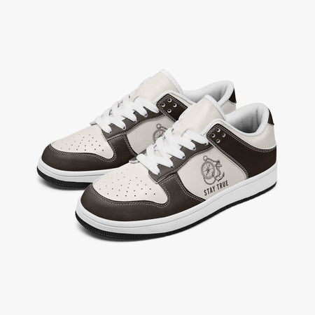 Angled three-quarter view of low-top sneakers in espresso brown and cream leather, featuring an anchor and compass graphic with "STAY TRUE", white laces, and a white inner lining.