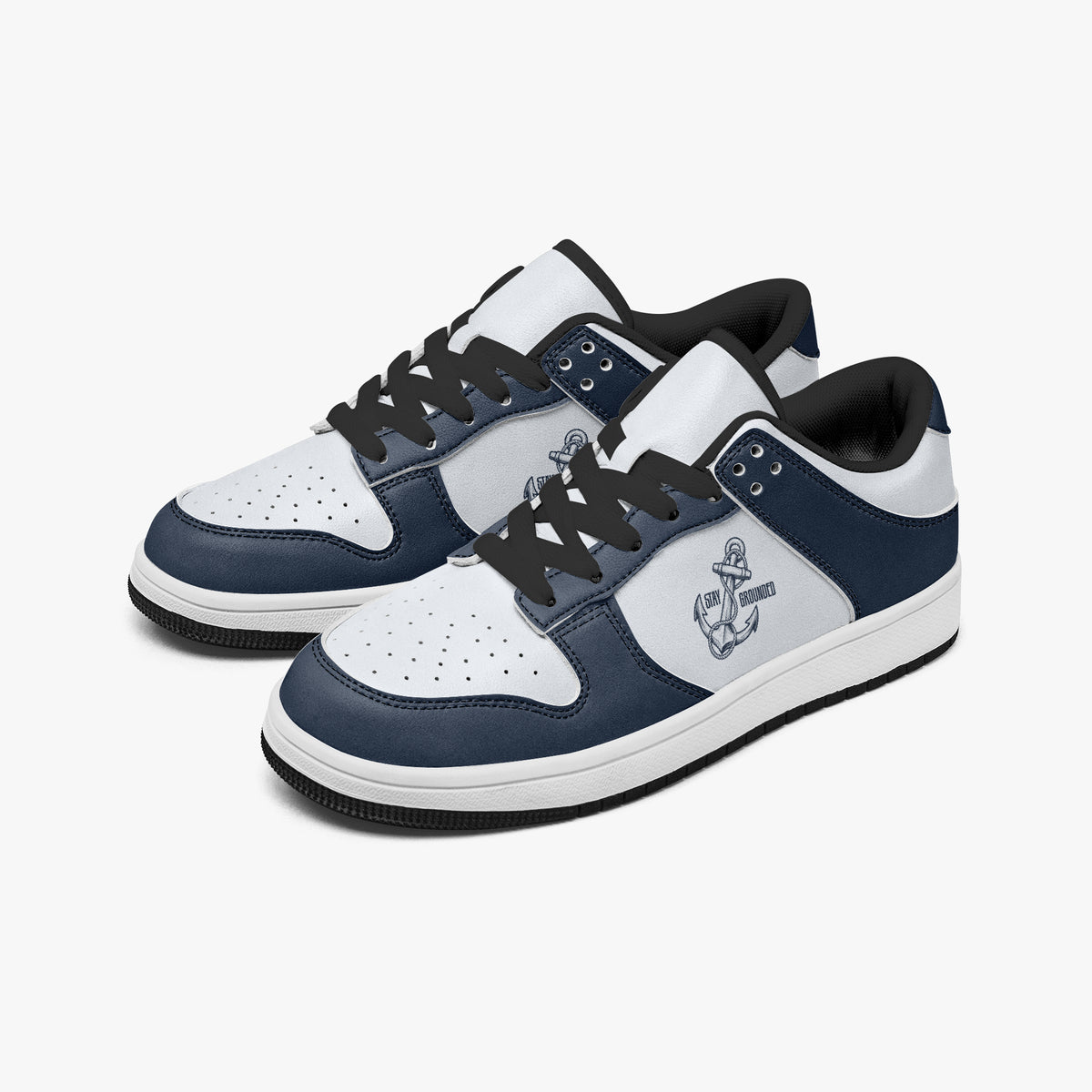 Angled three-quarter view of low-top sneakers in navy blue and white leather, featuring a maritime anchor graphic with "STAY GROUNDED" and black laces.