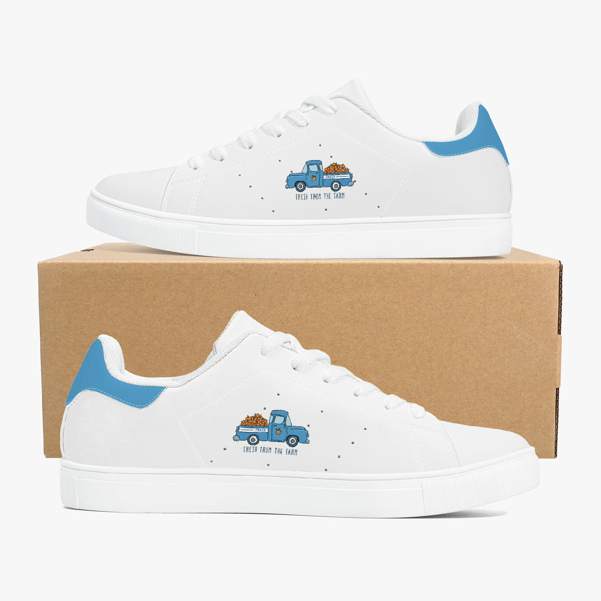 A pair of white low-top sneakers with a blue and brown cartoon pickup truck graphic on the side, displayed on and in front of a brown cardboard box against a light gray background.