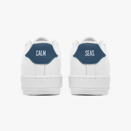 Rear view of white low-top sneakers showing contrasting navy blue heel tabs labeled "CALM" and "SEAS" above solid white rubber soles.
