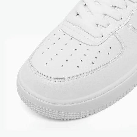 A close-up shot of a white low-top sneaker, showing the toe box with perforations, white laces, and a thick textured white sole on a clean white background.