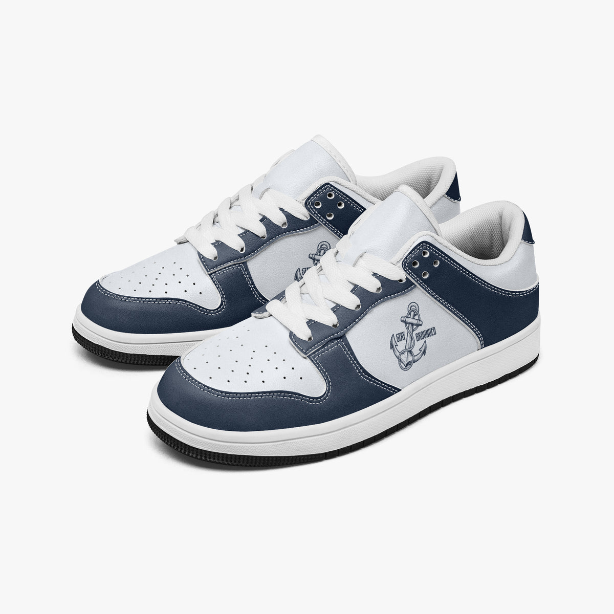 Angled three-quarter view of low-top sneakers in navy blue and white leather, featuring an anchor graphic with "STAY GROUNDED", white laces, and white inner lining.