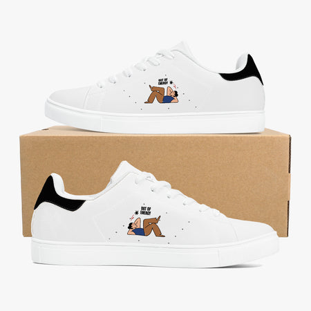A pair of white low-top sneakers with black heel tabs and a small cartoon graphic on the side, displayed on and in front of a brown cardboard shoebox against a light gray background.