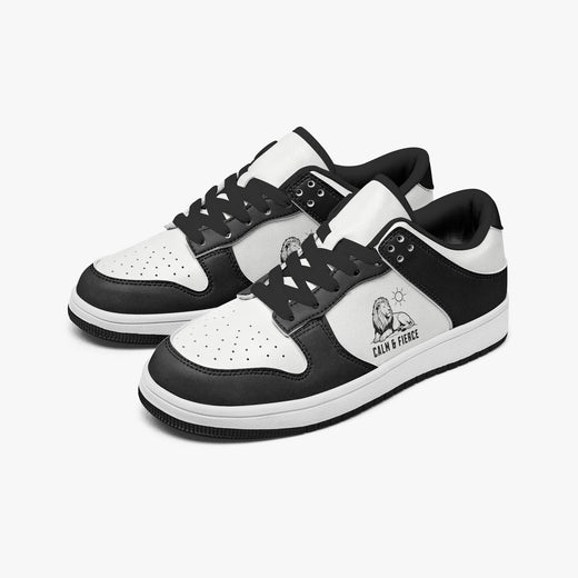 Angled three-quarter view of low-top sneakers in charcoal black and white leather, featuring a lion graphic with "CALM & FIERCE", black laces, and a black inner lining.