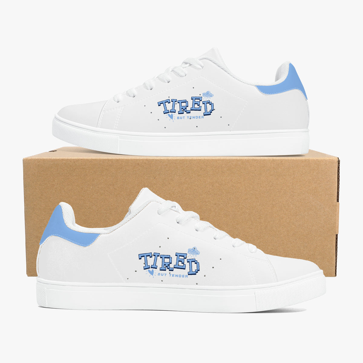 A pair of white low-top sneakers with light blue heel accents and a blue 'TIRED' graphic on the side, displayed on a brown cardboard box against a light gray background.