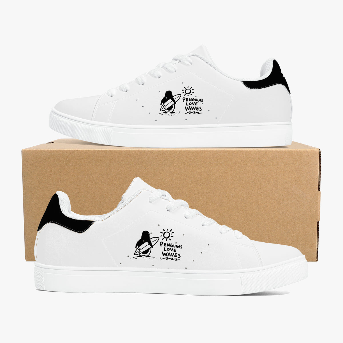 A pair of white low-top sneakers with black heel tabs and a black penguin graphic on the side that reads 'Penguins Love Waves'. The shoes are displayed on and in front of a brown cardboard box against a clean white background.